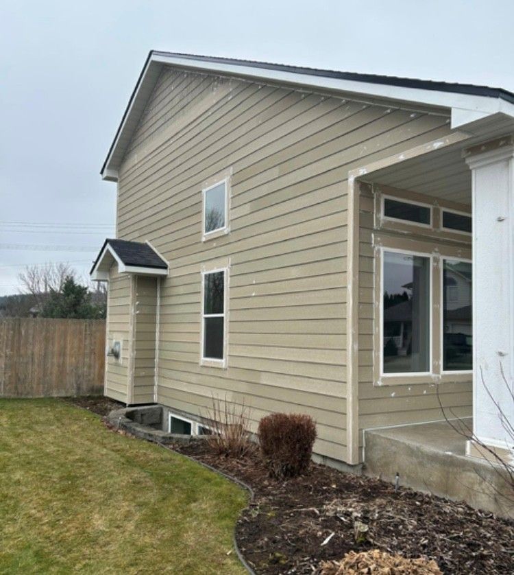 Beige house with light siding and dark roof, next to lawn and porch with large windows.