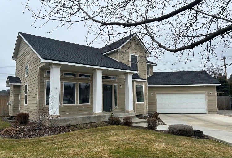 Beige two-story house with white pillars, black roof, attached garage, and driveway on a cloudy day.