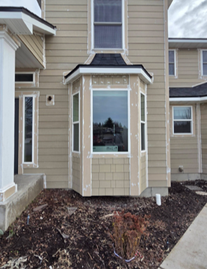 Beige house with a bay window, unfinished siding, and a patch of dirt in front.