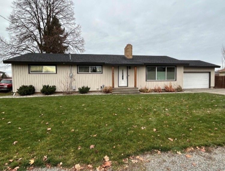 Beige ranch-style house with black roof, garage, and chimney, on a green lawn with shrubs under a cloudy sky.