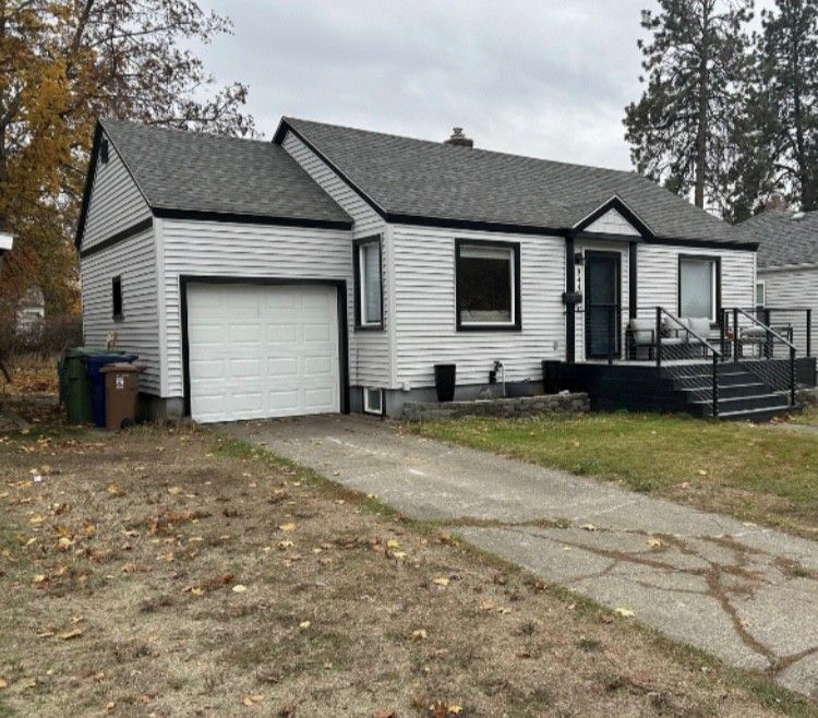 A one-story white house with black trim, a garage, and a cracked driveway on a cloudy day.