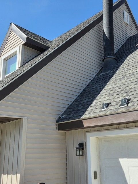 A low-angle view of a beige house exterior with a dark roof, brown trim, a chimney, a garage door, and a black light.
