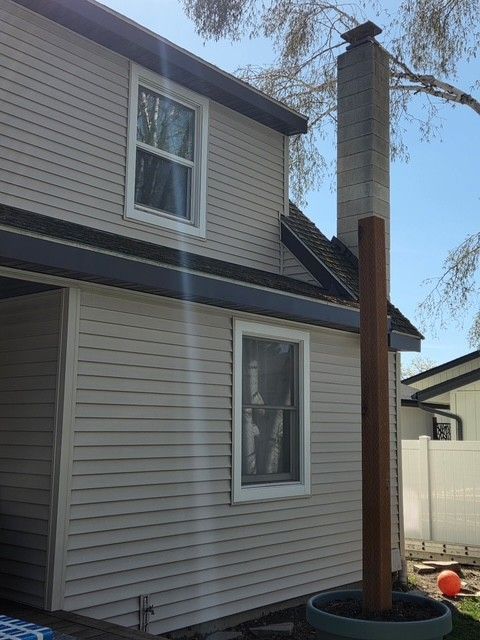 A light gray two-story house exterior with white-trimmed windows, a tall chimney, and a wooden post in a planter below.