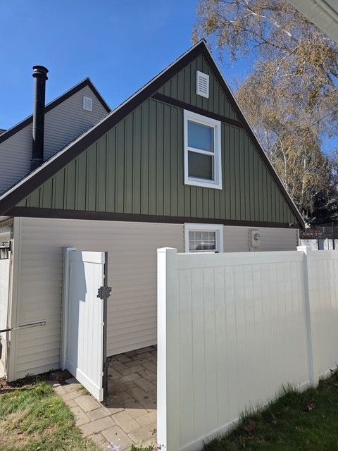 A two-story house with beige horizontal siding, a green-sided gable, brown trim, and a white vinyl fence with an open gate.