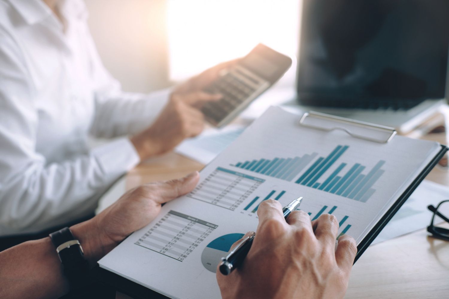 Dos personas analizan datos financieros de un informe con cuadros y gráficos, utilizando una calculadora y una computadora portátil.