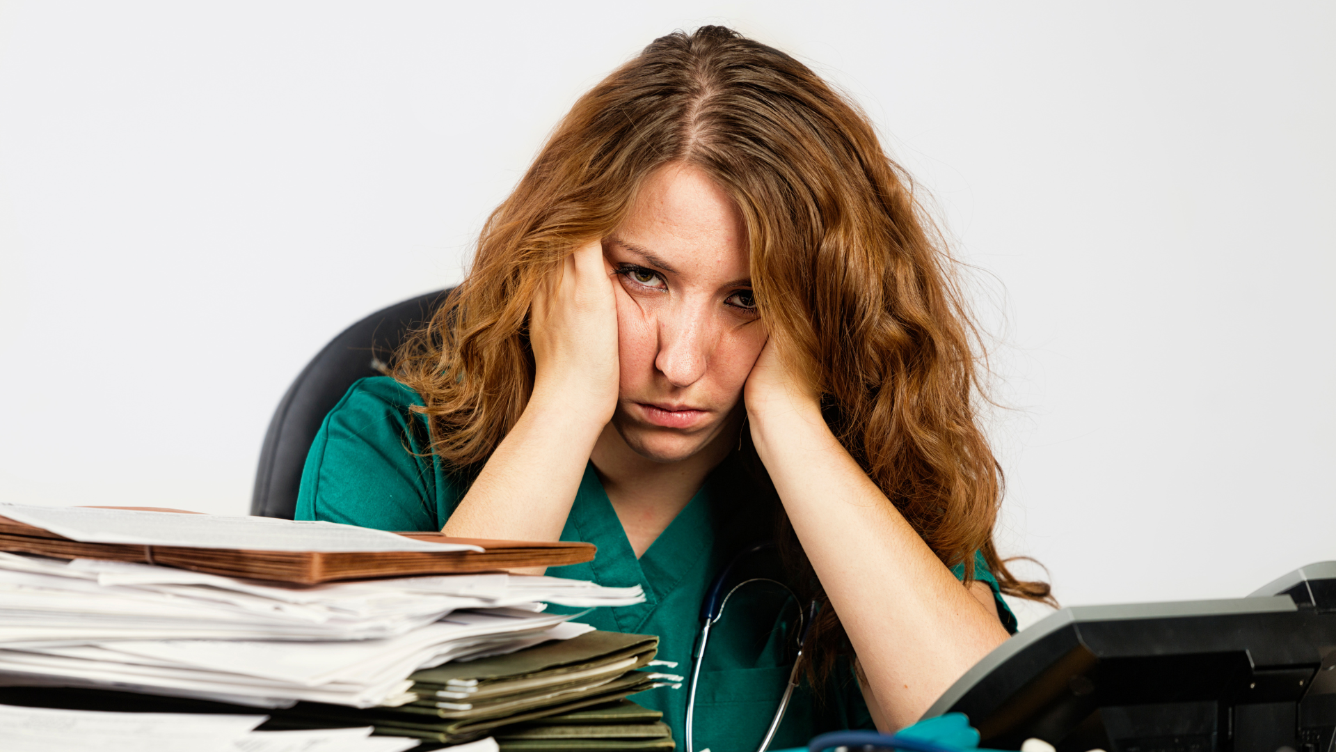 A stressed-out fertility care provider sits desk with her head resting on her hands, surrounded by paperwork.