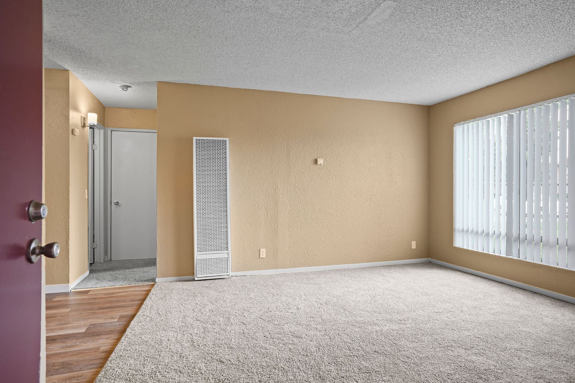 Empty beige carpeted living room with sliding glass door and vertical blinds, seen from the entryway