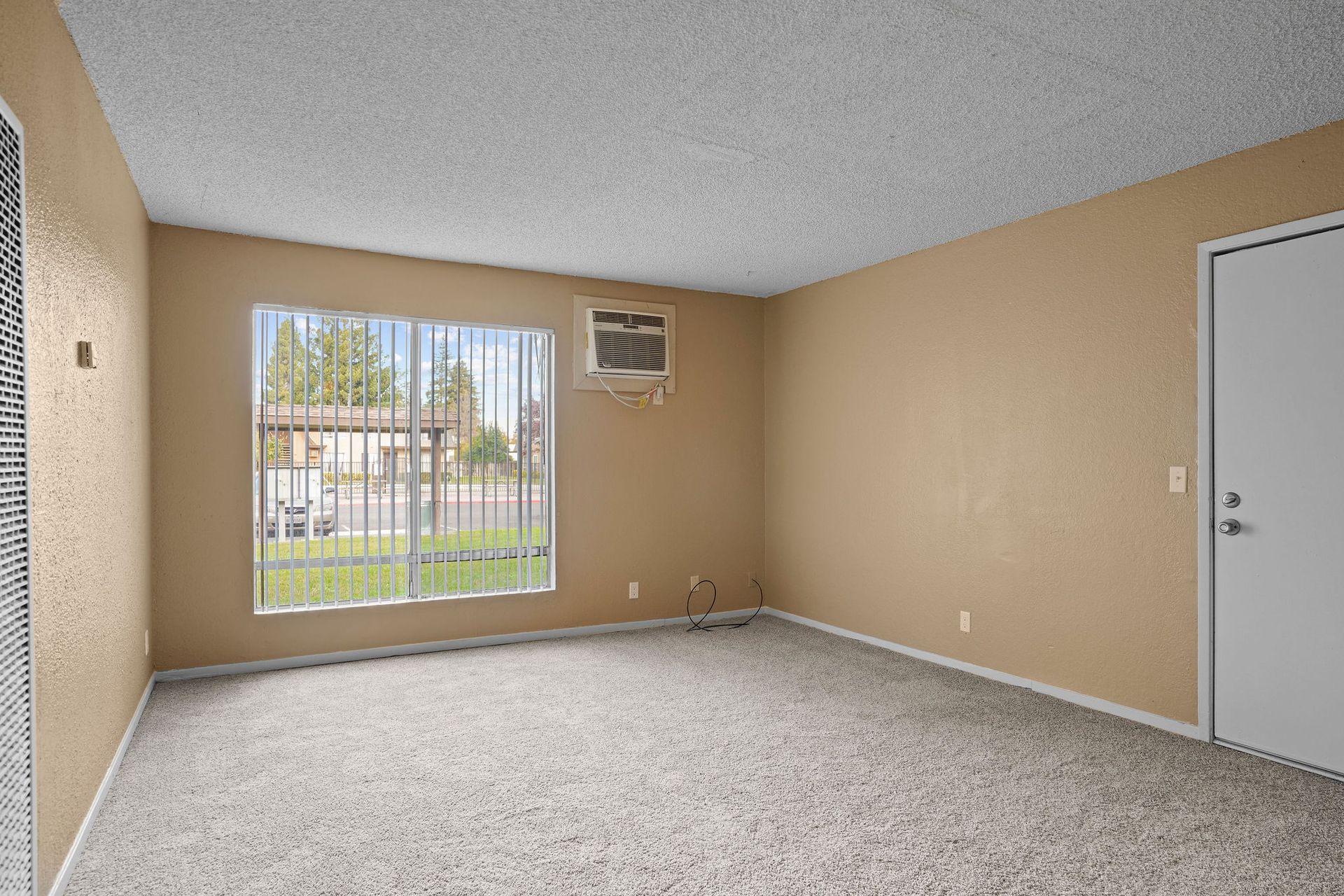 Empty beige carpeted room with large window, white door, and wall-mounted air conditioner.