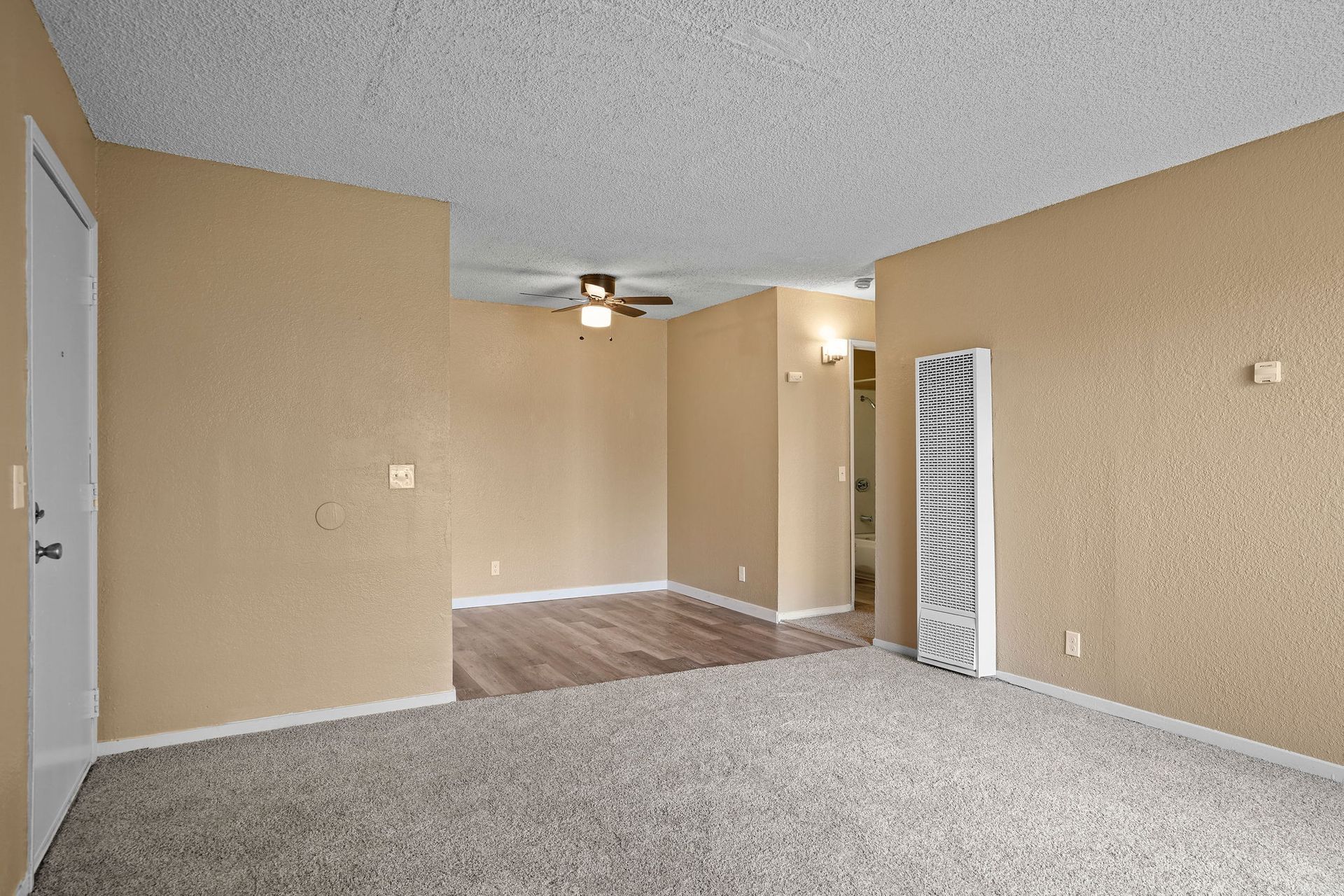 Empty beige living room with carpet, ceiling fan, and doorway to a wood-floored area
