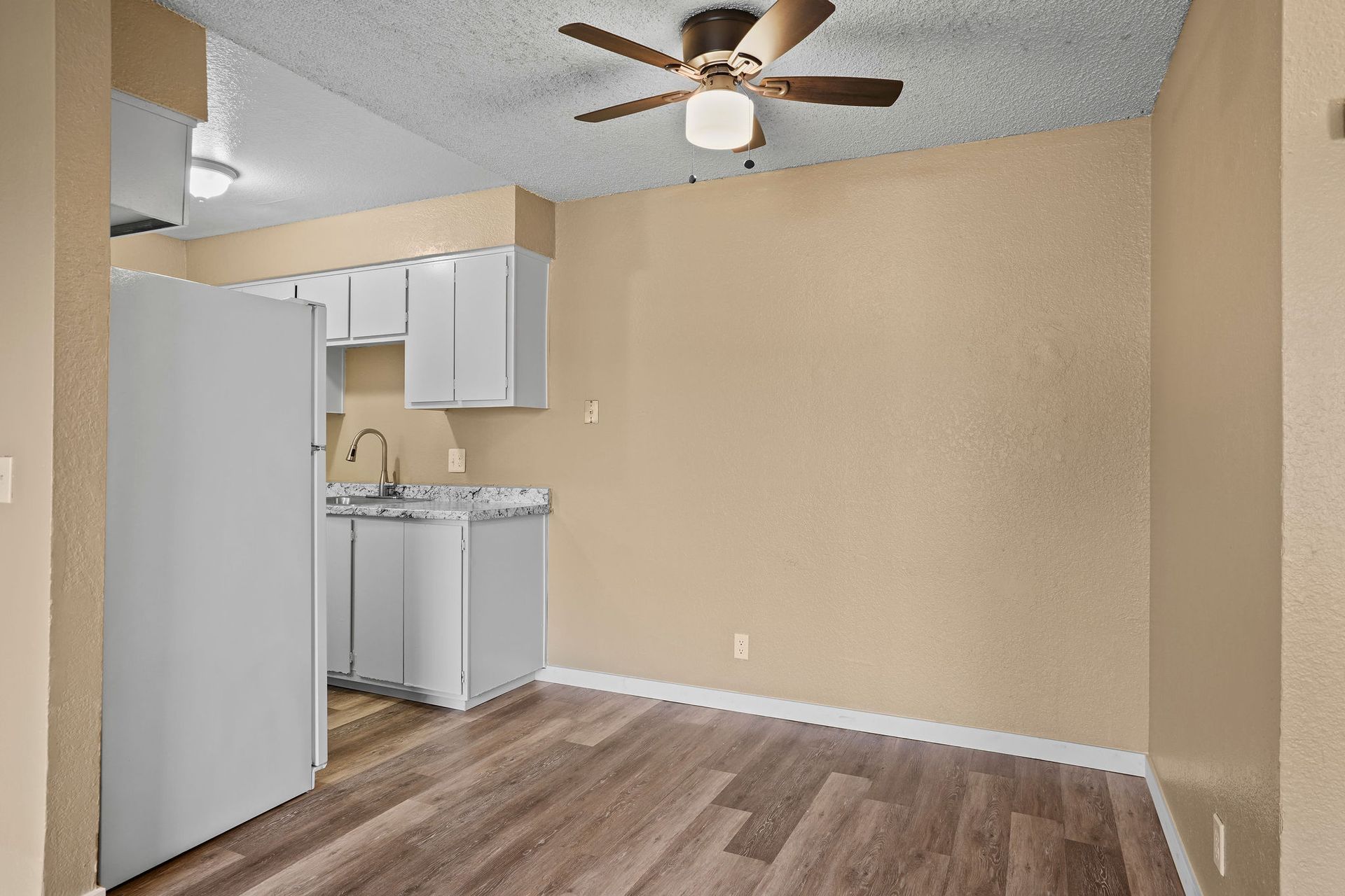 Empty beige living room with wood flooring, ceiling fan, and a small kitchen area with white cabinets.