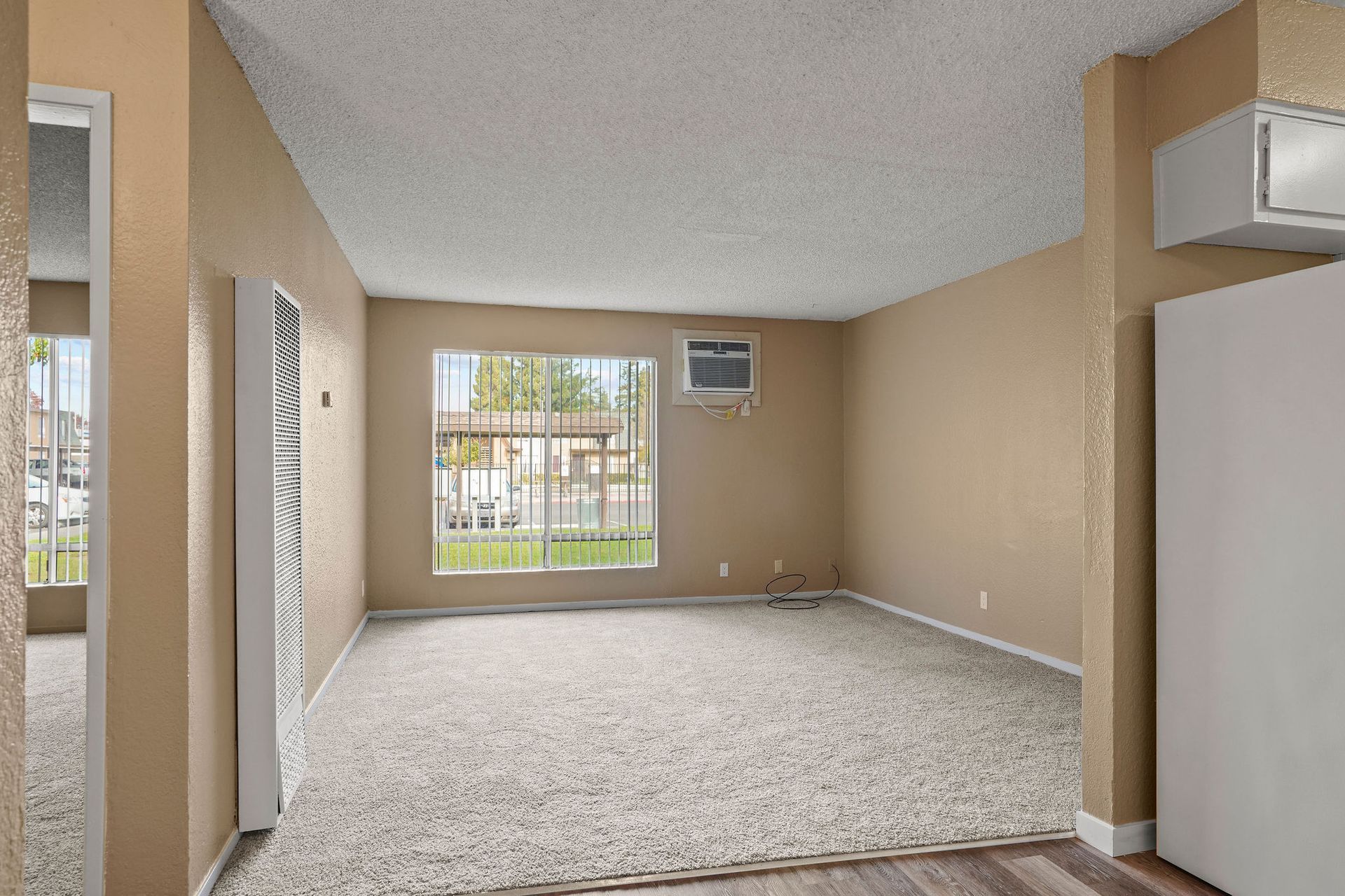 Empty beige living room with carpet, window, and wall-mounted air conditioner