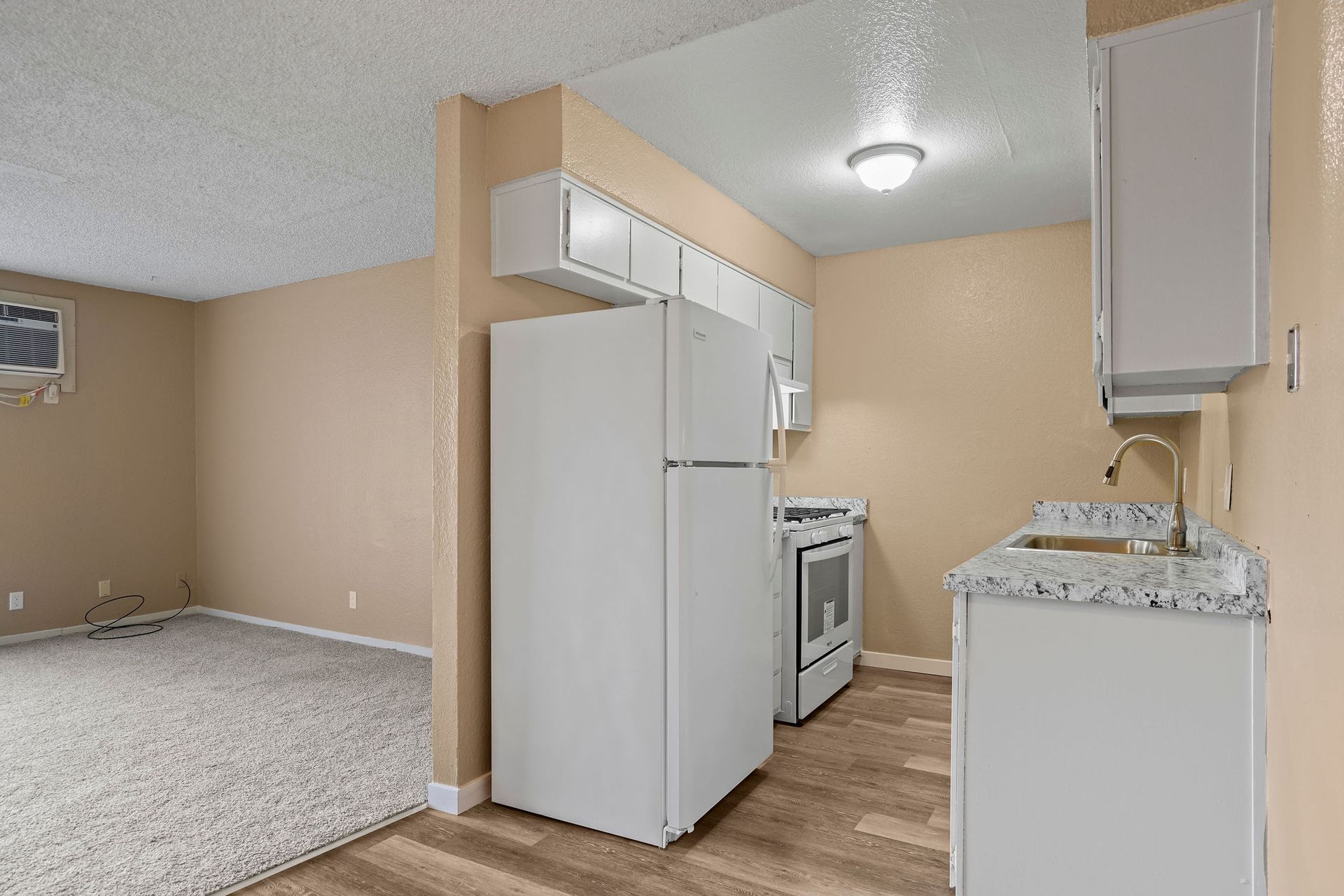 Empty apartment kitchen with white appliances, beige walls, and wood flooring leading to a living area