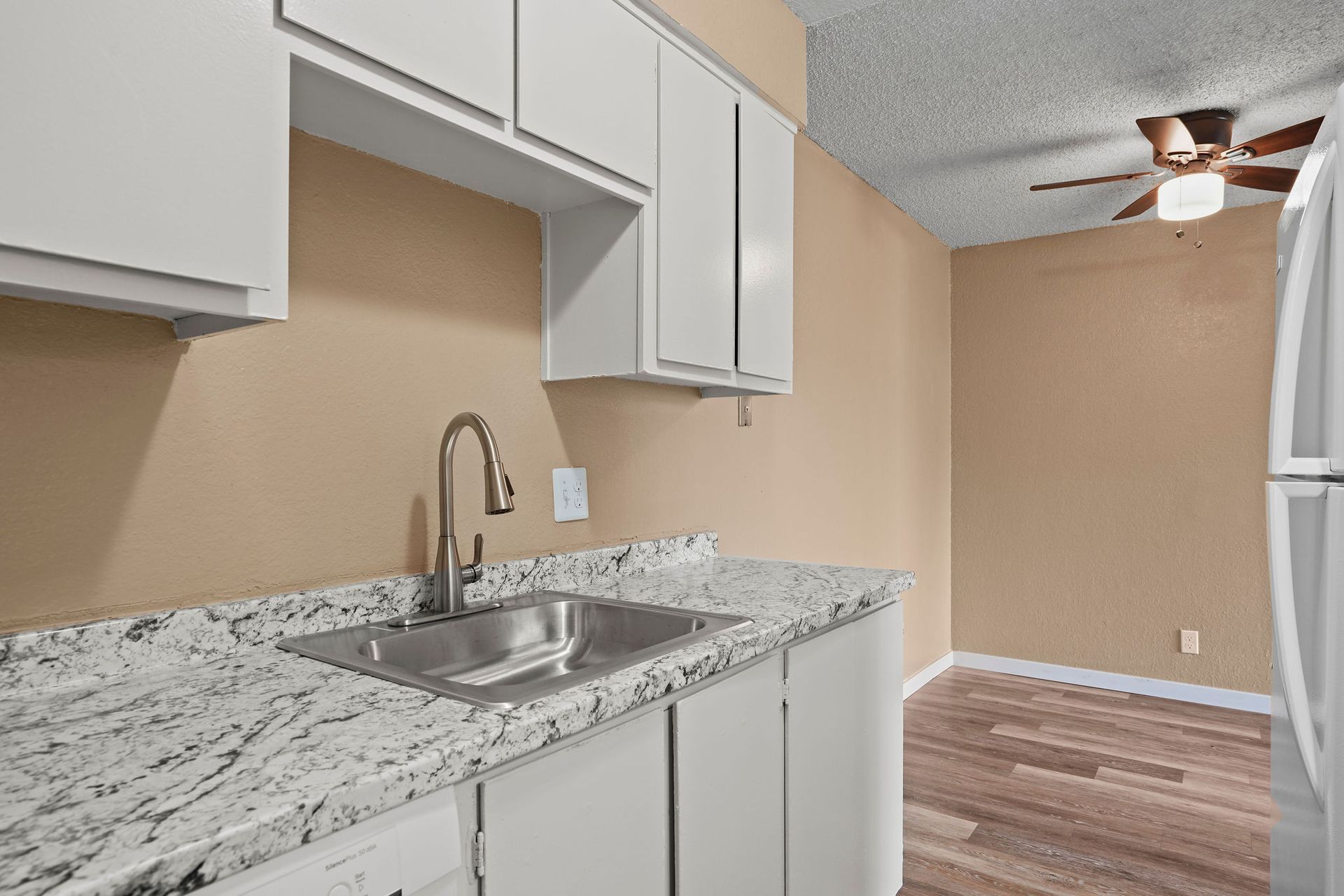Small kitchen with white cabinets, granite countertop, stainless sink, and beige walls opening to a dining area.