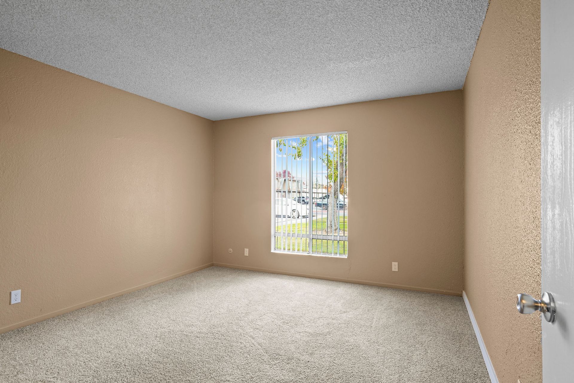 Empty beige bedroom with carpet and a small window overlooking houses outside.