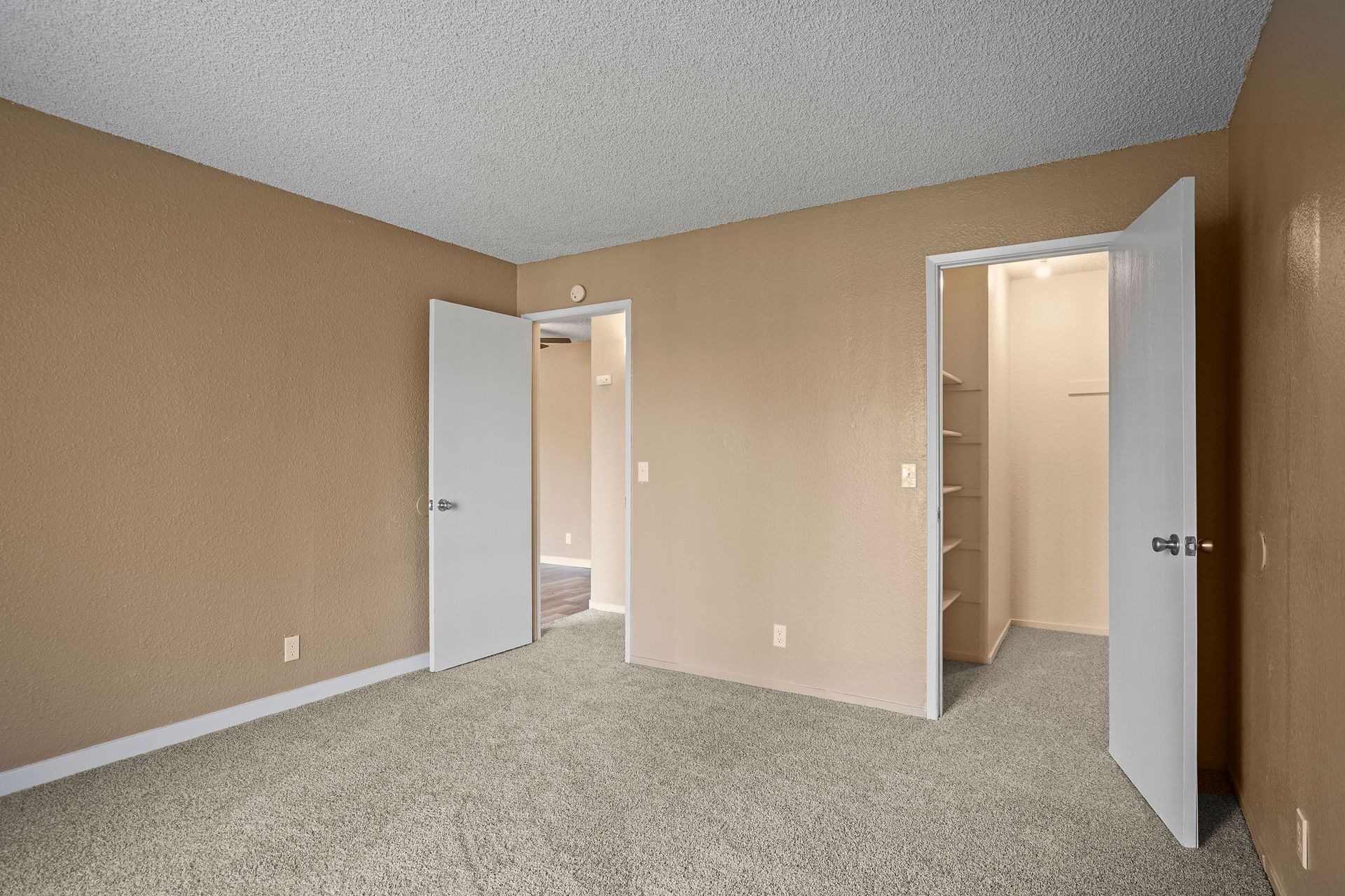 Empty beige bedroom with carpet and two open doors leading to a lit closet and hallway