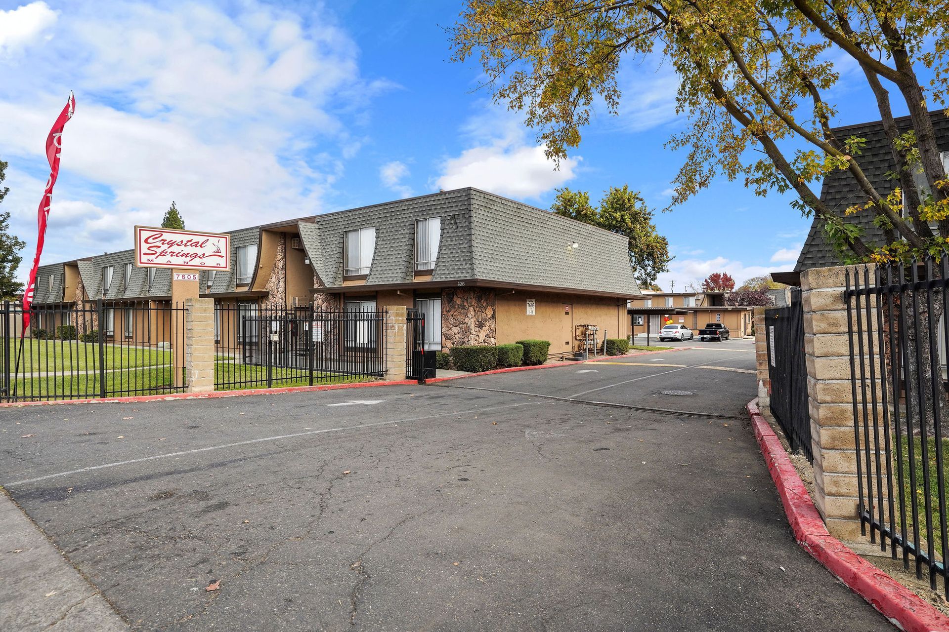 Motel-style building with a paved driveway, black fence, and autumn trees under a blue sky.