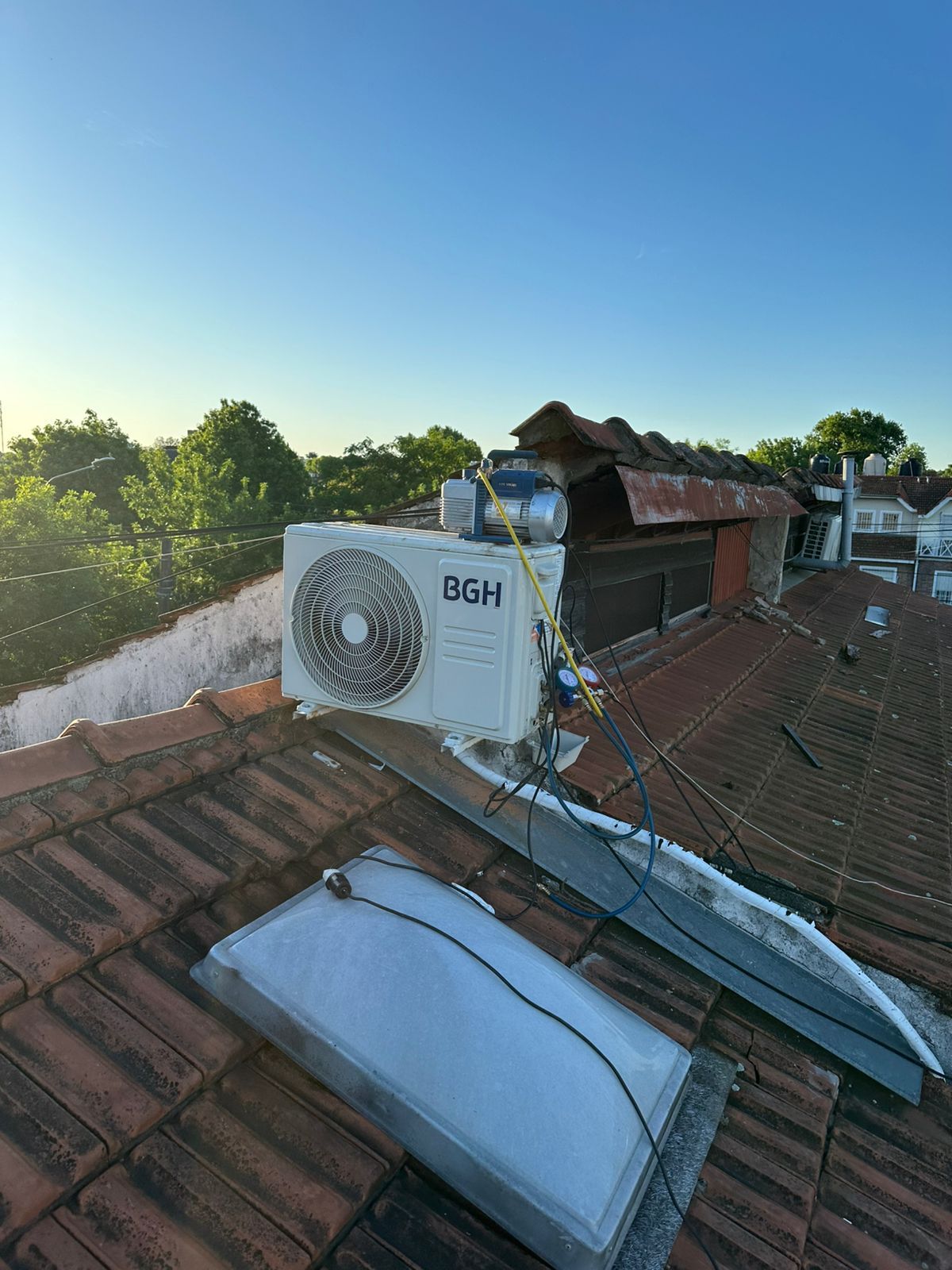 Unidad de aire acondicionado en un tejado de tejas con tragaluz; cielo azul al fondo.