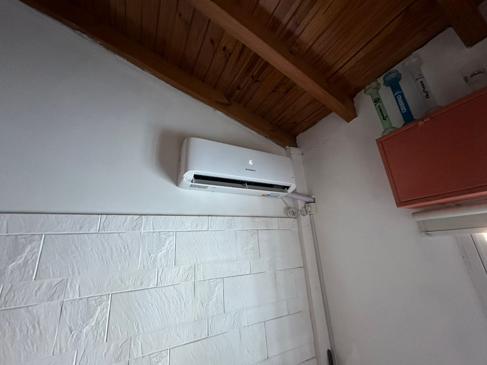 Aire acondicionado blanco sobre una pared blanca texturizada, techo de madera. Armario rojo visible a la derecha.