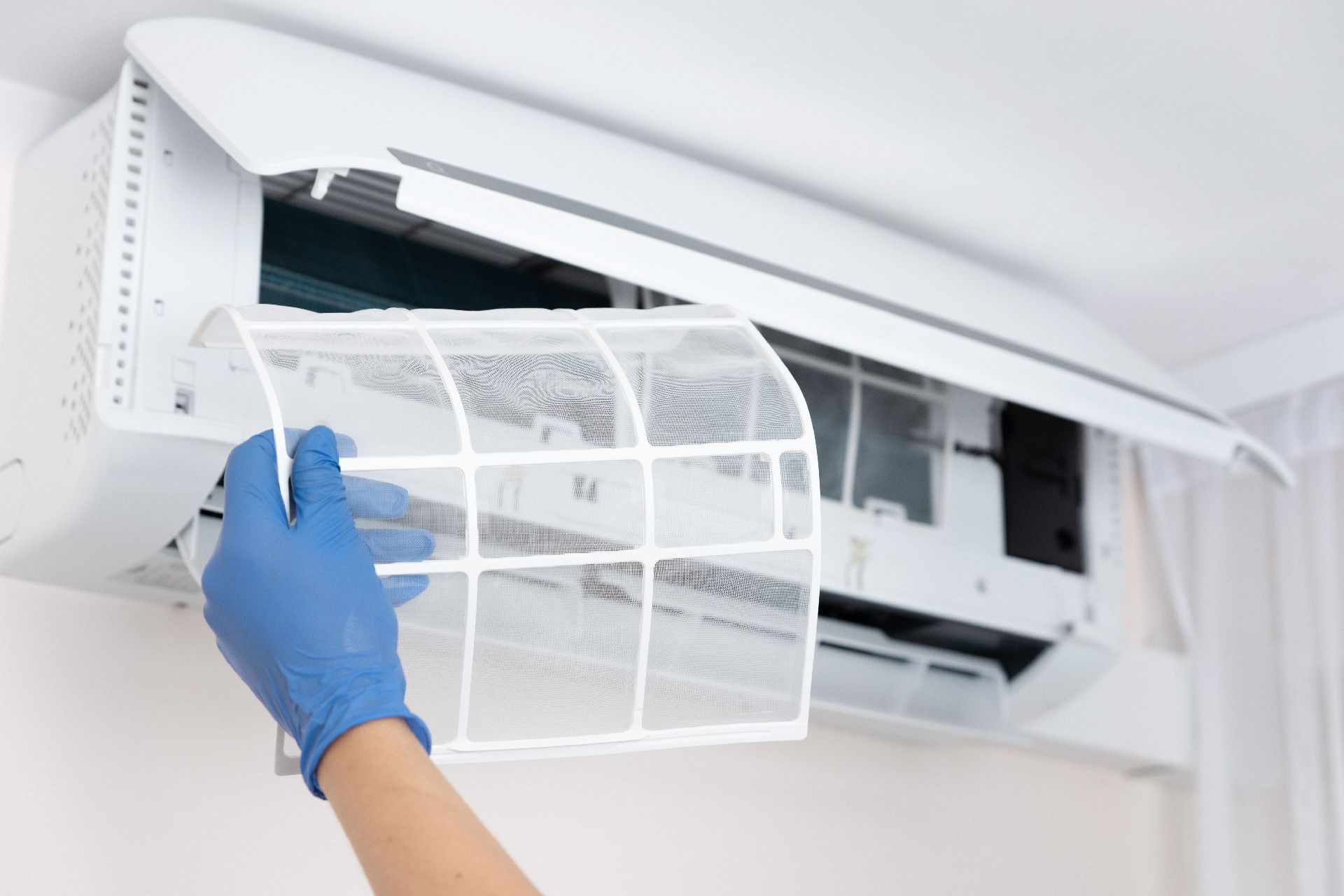 Persona con guantes azules retirando un filtro de aire acondicionado. Unidad blanca en la pared.