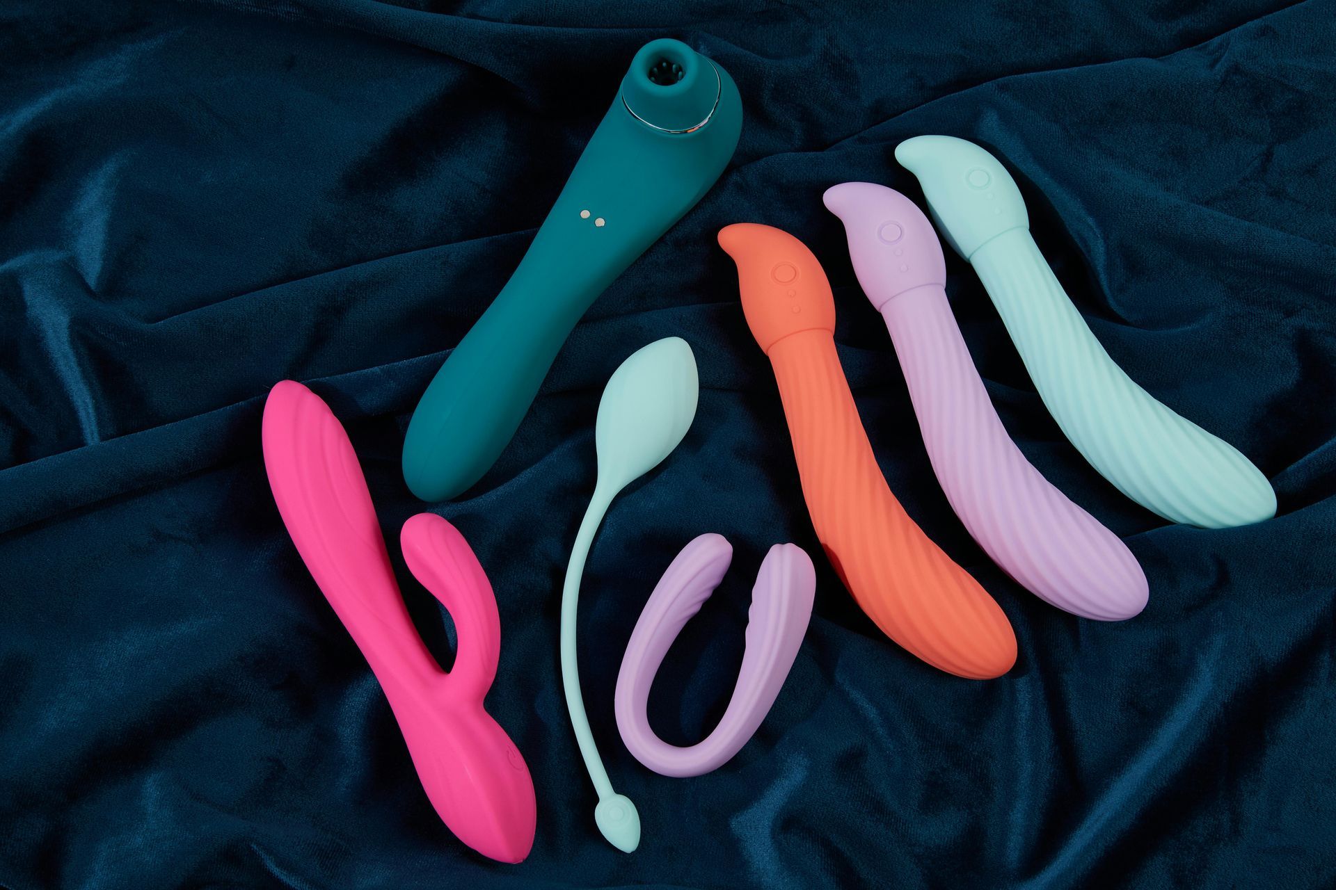 A collection of various vibrators in pink, orange, teal, and light blue arranged on a dark blue fabric background.
