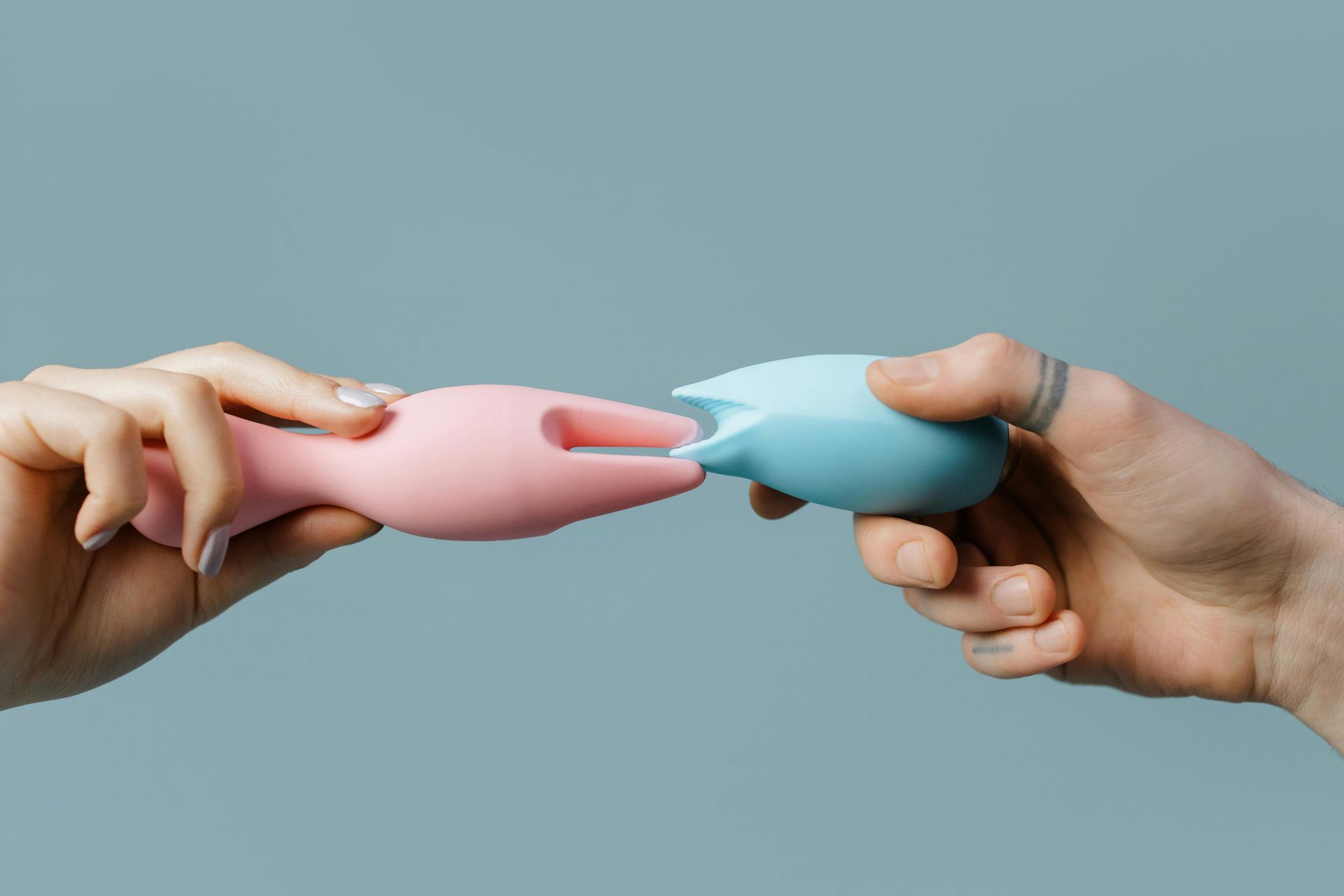 Two hands hold a pink and a blue silicone object against a solid light blue background, connecting them at the center.