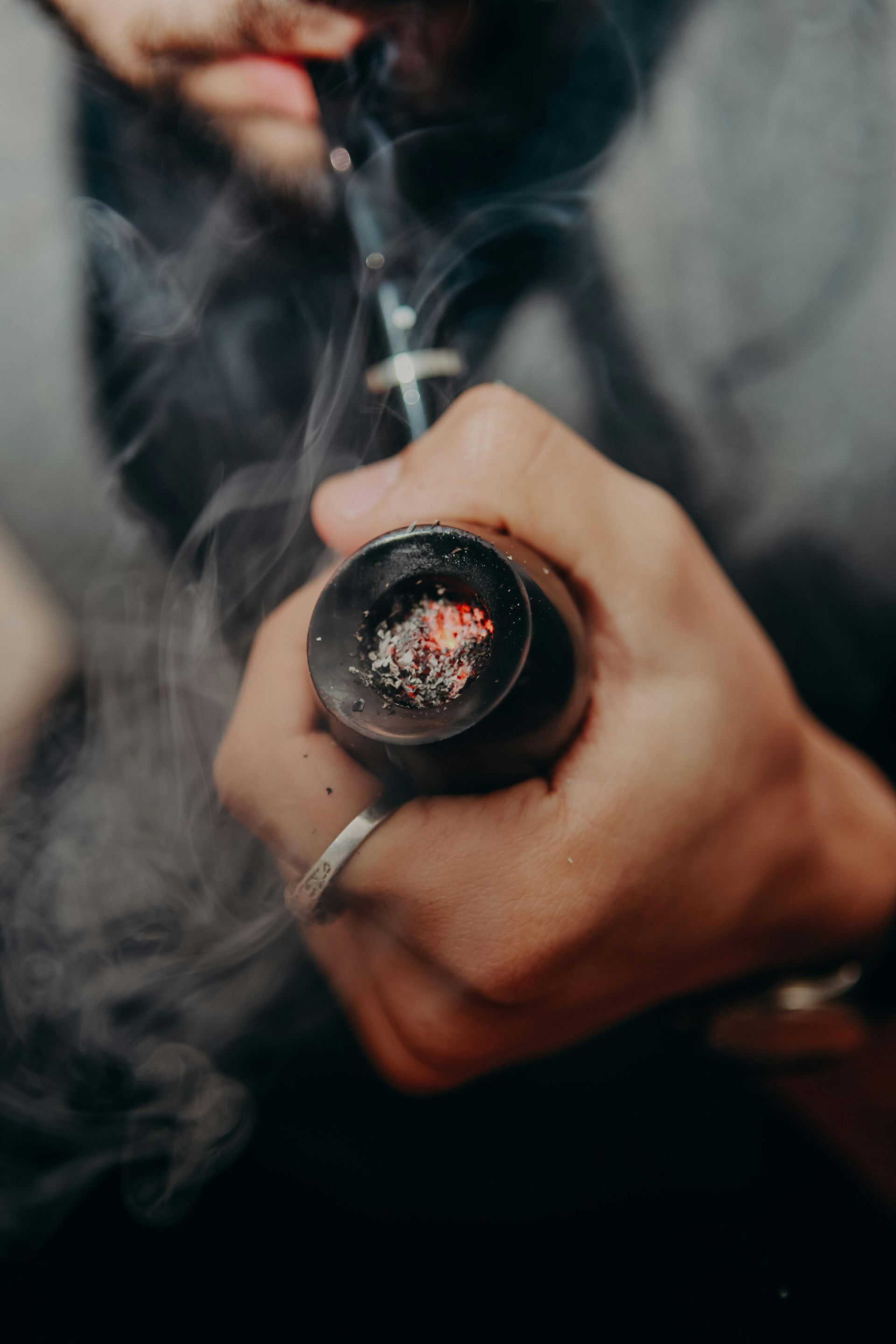 A close-up view of a hand holding a smoking pipe with glowing embers inside, surrounded by wisps of grey smoke.