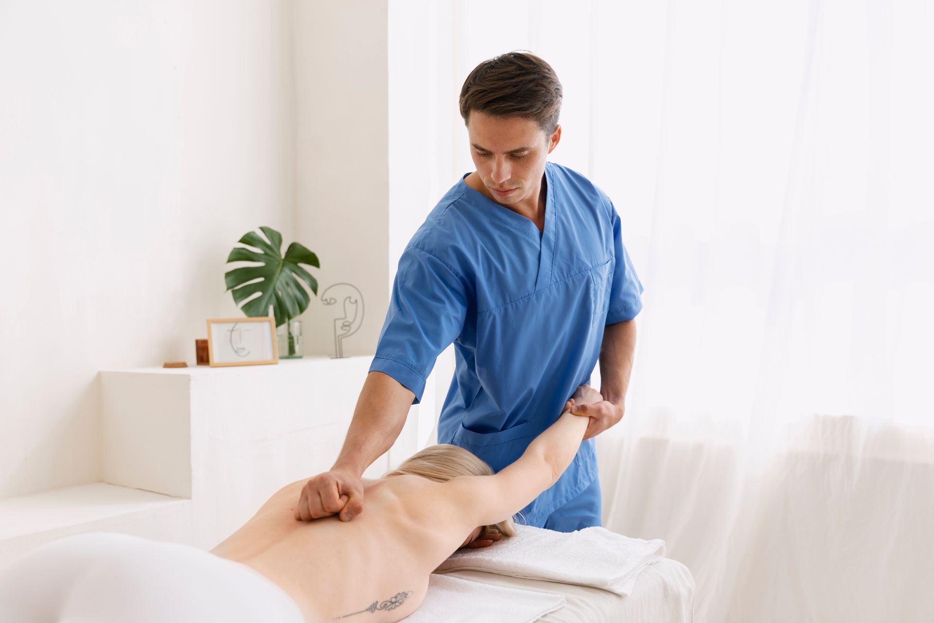 A professional in blue scrubs performs a massage on a client's back in a bright, minimalist room.