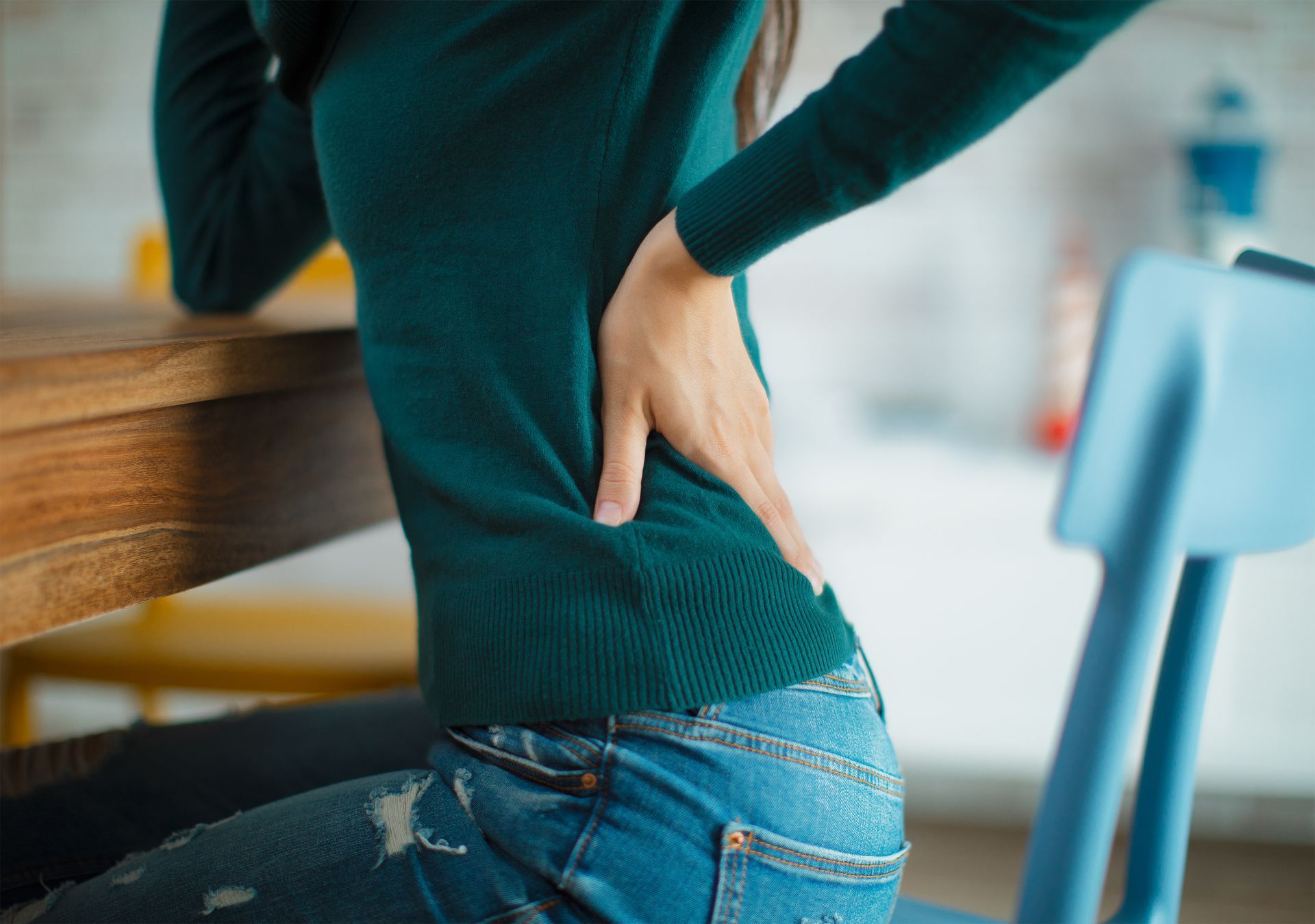 A person in a green sweater and jeans sitting at a table, holding their lower back with their hand.