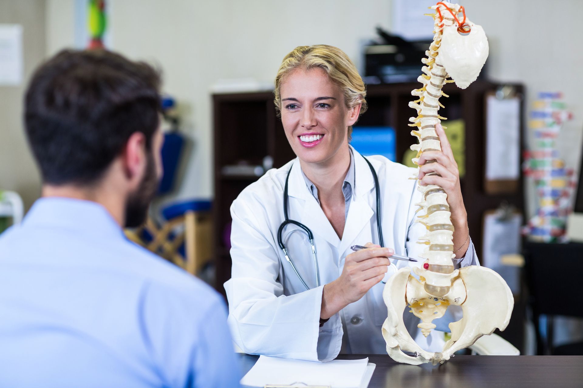 Doctor pointing at a spine model while speaking to a patient in a doctor's office.