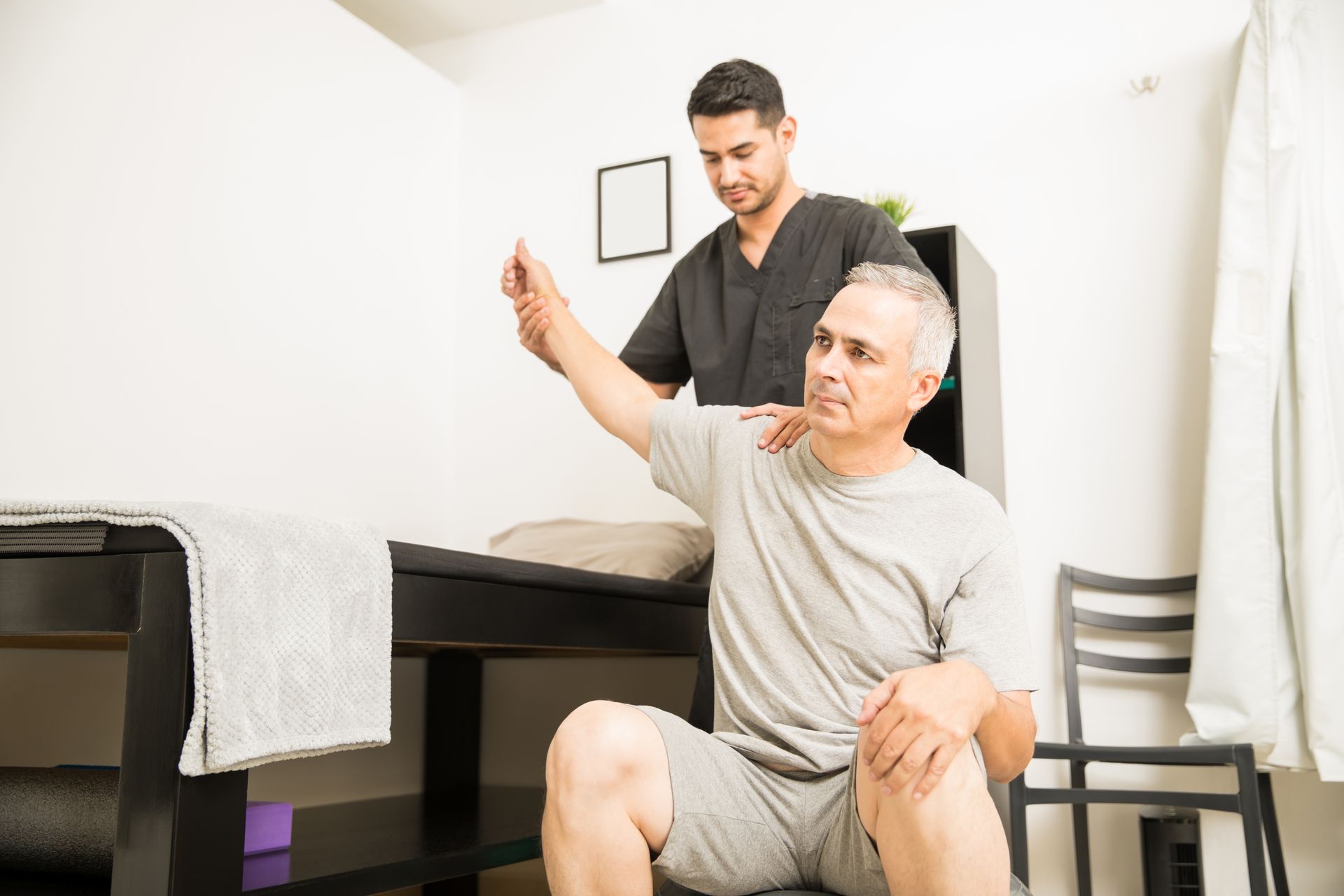 A physical therapist assists a patient with a seated shoulder stretch in a clinical office.