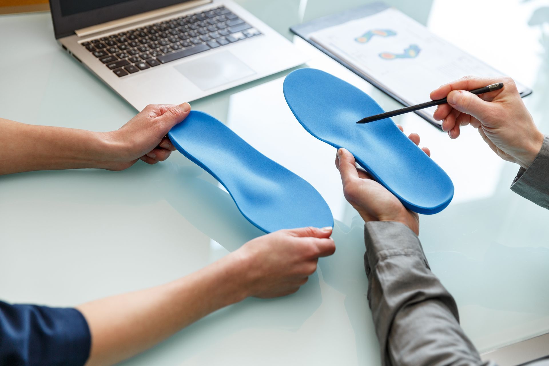 Two people hold and discuss custom blue shoe insoles over a desk with a laptop and a document.