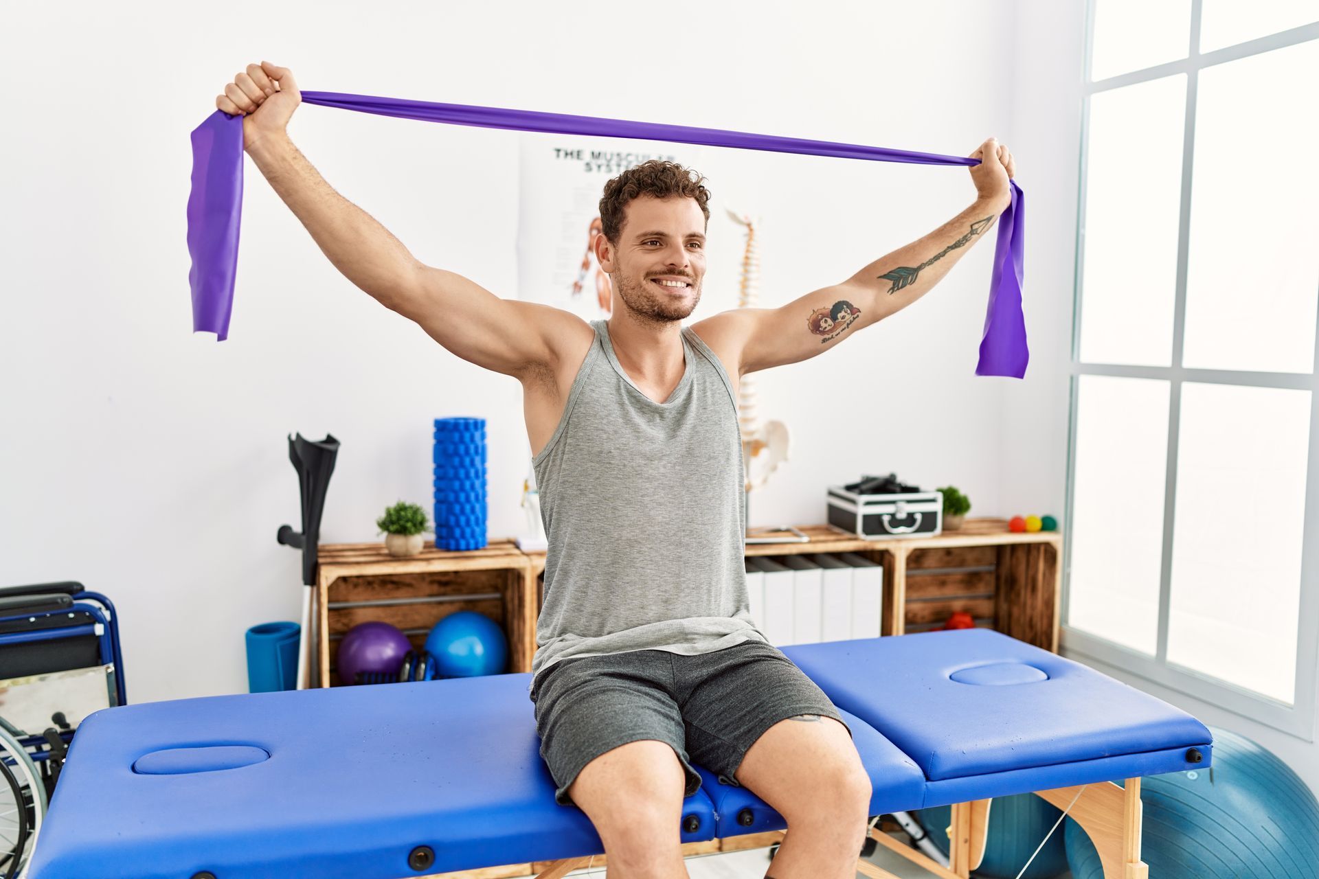 A man is sitting on a blue table holding a purple exercise band.