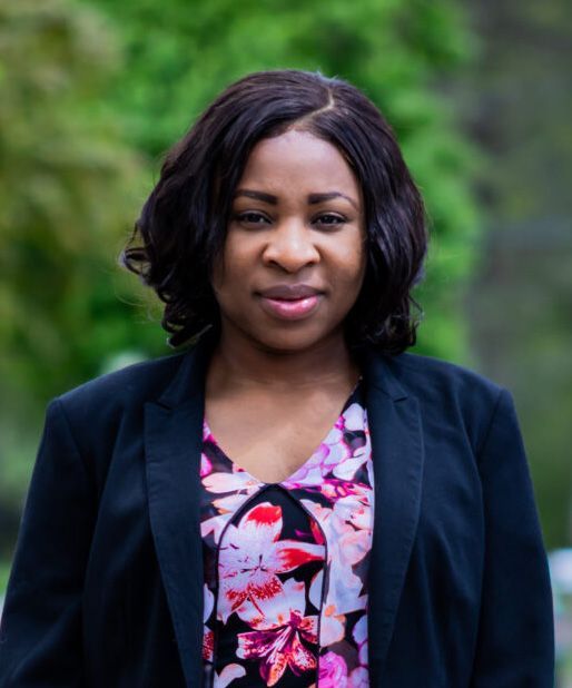 Woman with dark hair in black blazer, floral shirt, standing outside.