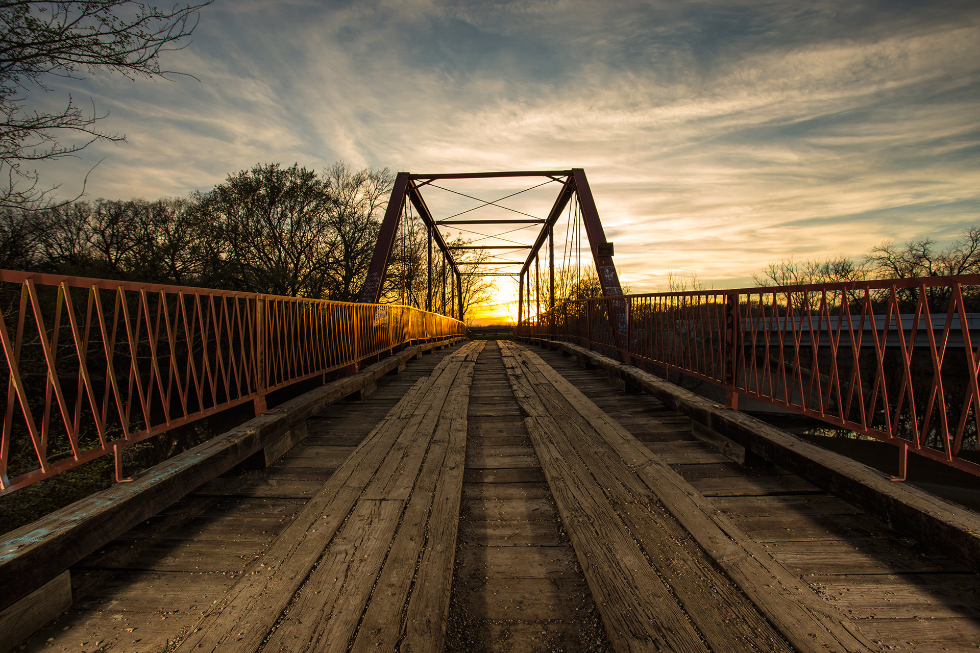 Wooden bridge at sunset with orange railings and a metal arch; golden sky in background.