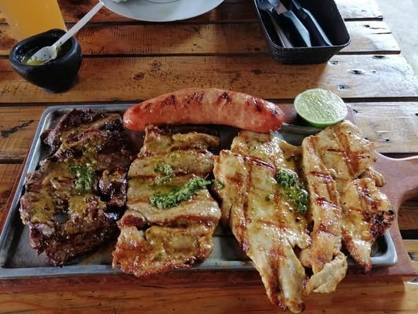 Plato de carne a la parrilla con salchicha, lima y salsa sobre una mesa de madera.