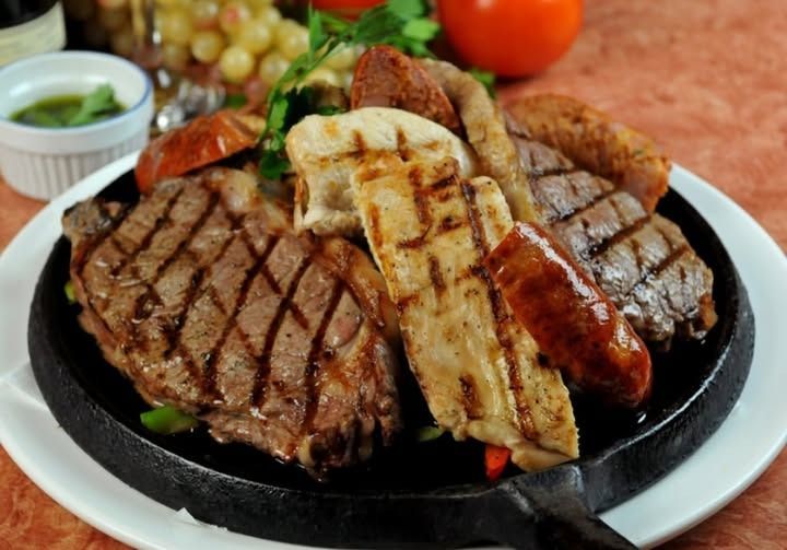 Carnes a la parrilla: bistec, pollo, salchicha y pan en una sartén caliente, adornado con hierbas y salsa.