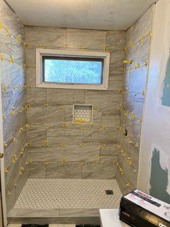 Shower tiled with gray rectangular tiles, window, recessed shelf, and small white hexagonal floor tiles.