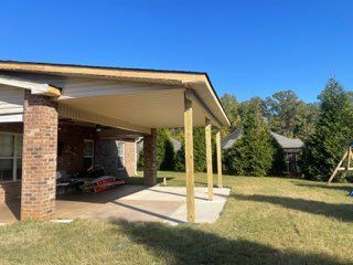 A brick house with a covered patio, wooden beams, and a concrete floor on a green lawn. Blue sky.