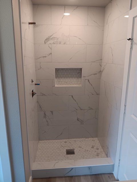 Shower with marble-look tiles, small recessed shelf, and patterned floor. Open doorway to the right.