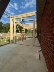 A wooden pergola under construction, attached to a brick building; concrete patio.