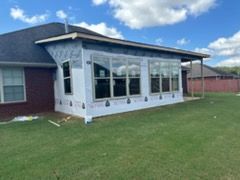 Backyard of a house with an addition under construction, windows installed, wrapped in a blue material, with a green lawn and blue sky.