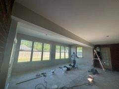 Workers inside a room, one on a ladder, others on the floor with tools, unfinished drywall, light gray walls, large windows.