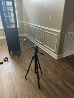 A laser level on a tripod projecting a green line for installing wall trim in a room with wood flooring.