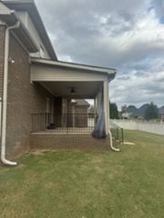 A brick house with a covered porch, brown railing, and grassy yard under a cloudy sky.