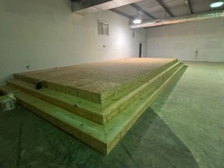 Wooden tiered stage in a large, empty room with concrete floor and white walls.