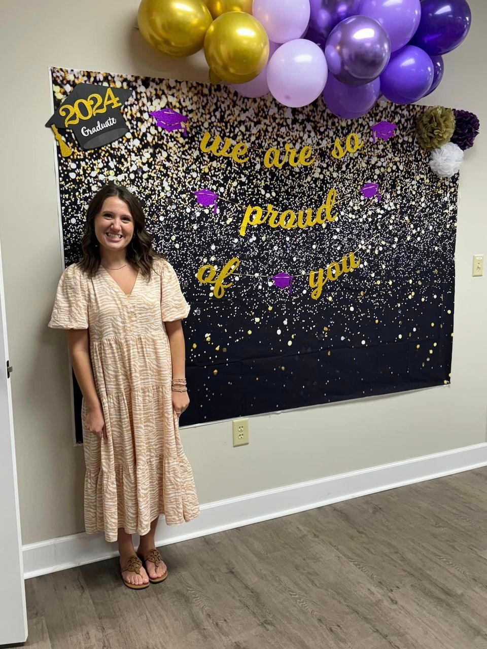 A woman in a dress is standing in front of a sign that says we are so proud of you.
