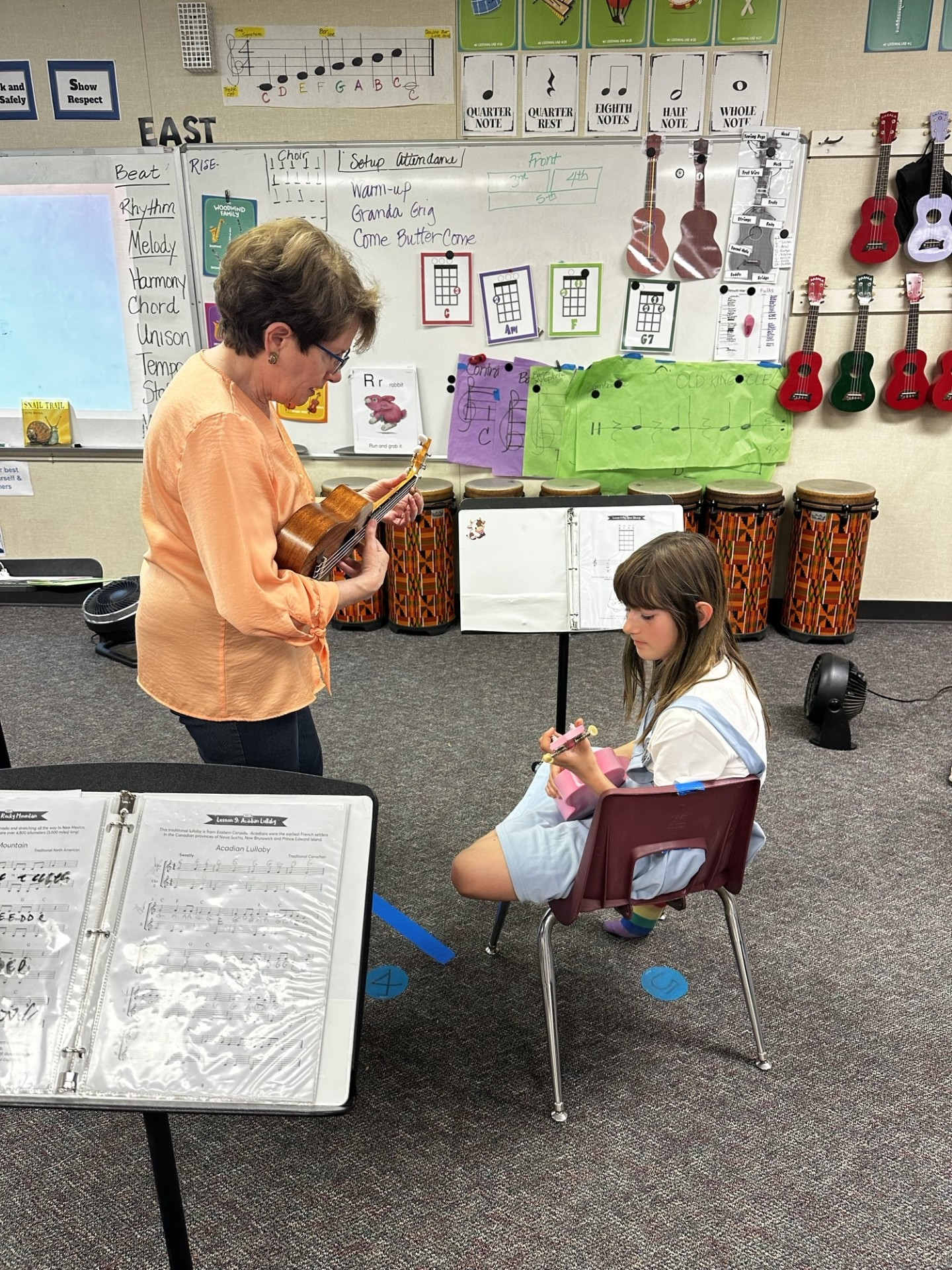 A woman is teaching a young girl how to play an ukulele in a classroom.