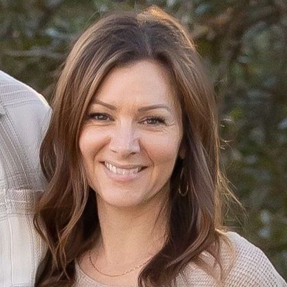 A woman with long brown hair is smiling in front of a body of water.