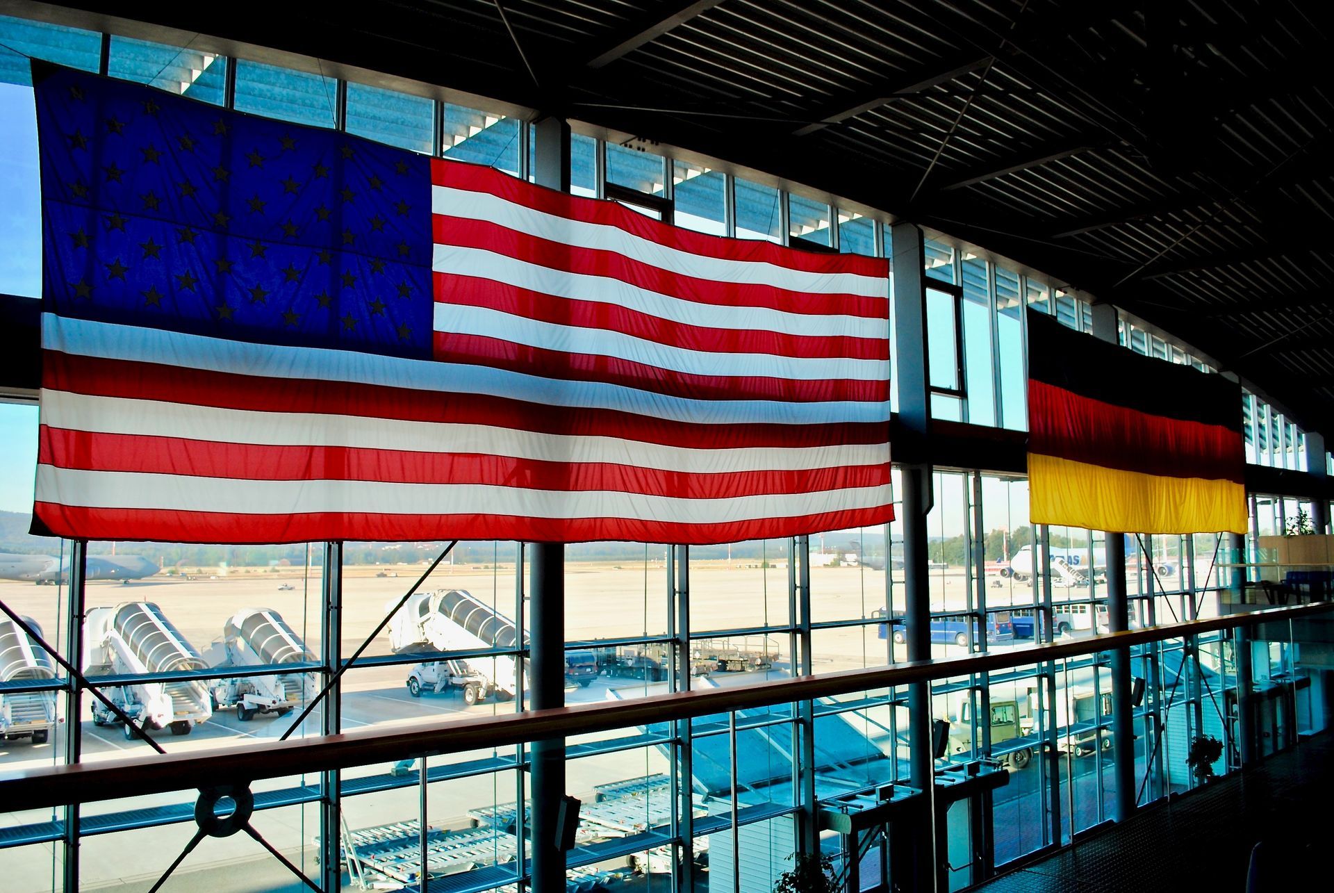 American and German flags hanging inside an airport, with a view of the tarmac outside.