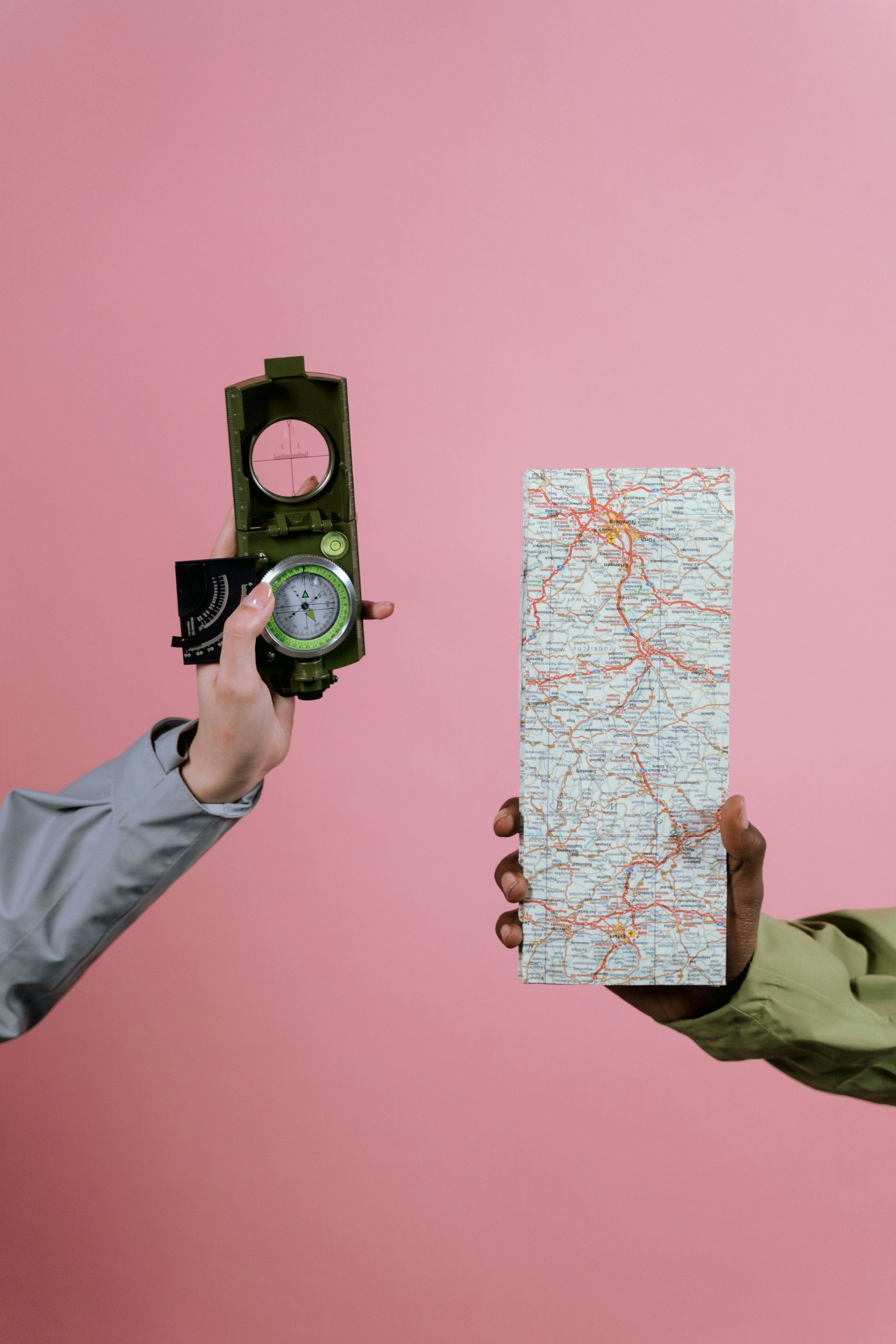 A picture of a compass and a map with a womans finger in shot holding the compass on a block coloured watermelon pink background
